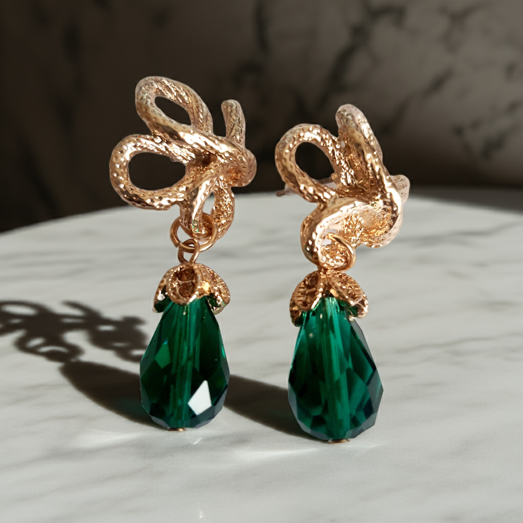 Złote kolczyki ze stali nierdzewnej w stylu glamour, ozdobione rzeźbioną kokardą i zielonym kryształem w kształcie kropli. Elegancja i szmaragdowy blask. Długość 6 cm. Rouge Bizu.