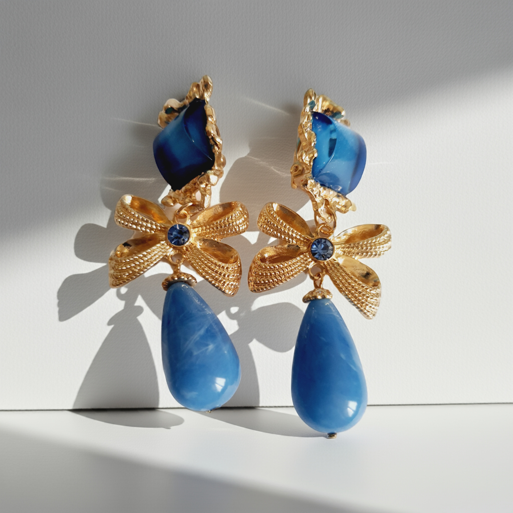 Złote klipsy ze stali nierdzewnej z niebieskimi kryształami. Dekoracyjne złote kokardy z błękitnym kamieniem i dużą kroplą w odcieniu niebieskiego. Styl glamour, romantyczna elegancja. Długość 7 cm. Rouge Bizu.