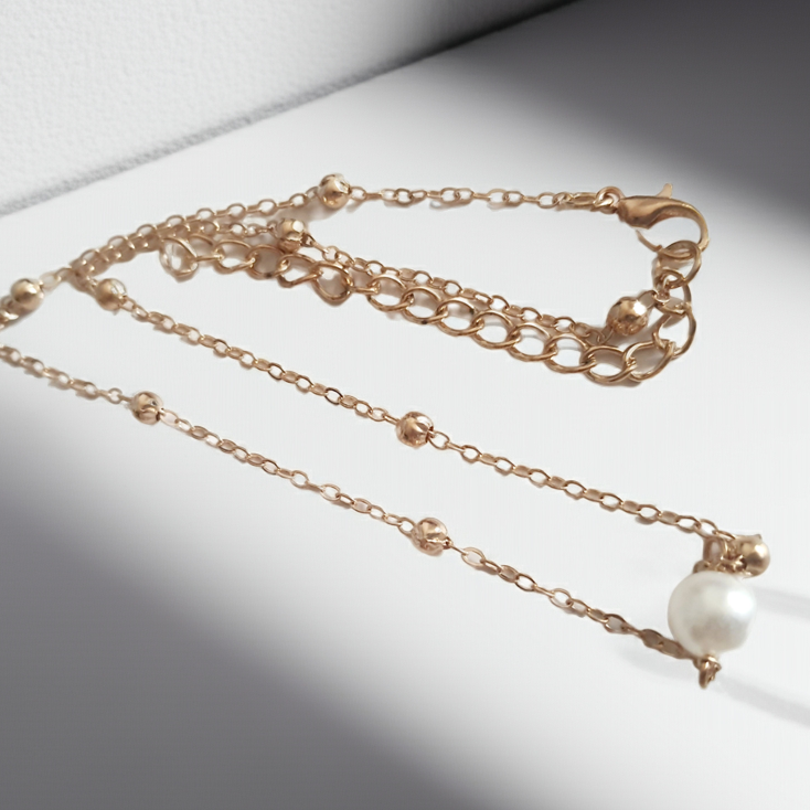 Złoty naszyjnik ze stali nierdzewnej z kulkami i zawieszką z białą perłą, ułożony na jasnym tle, typ celebrytka
ROUGEBIZU